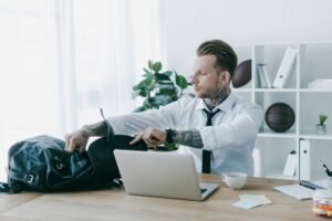 young tattooed businessman opening backpack while working with laptop at workplace