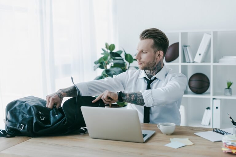 young tattooed businessman opening backpack while working with laptop at workplace