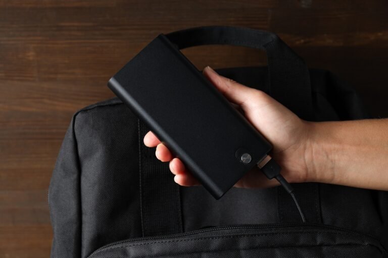 Powerbank in hand and backpack on wooden background, top view